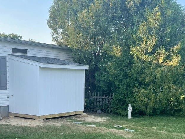 A small lean-to addition on the back of a single-story house: white vertical siding, a shed roof with gray shingles and a door on the house side, seen from the yard.