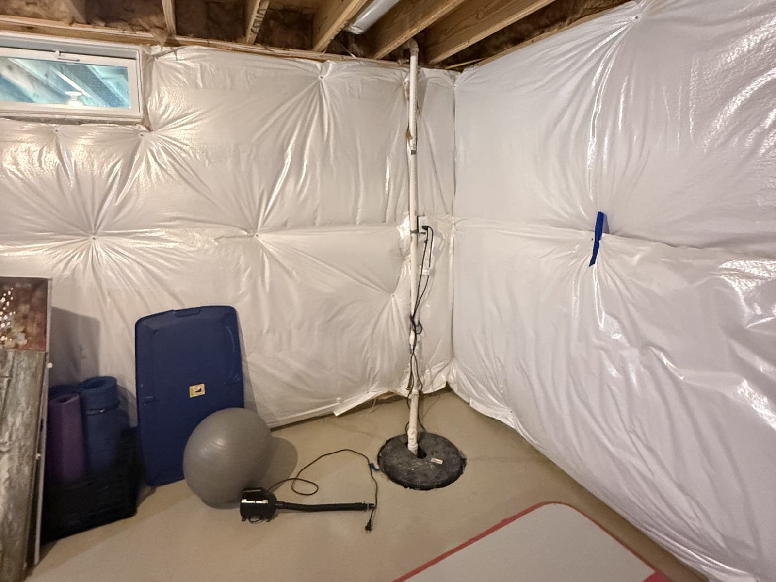 An unfinished basement corner: insulation blankets on the foundation walls, a small window, a sump pit with its discharge pipe, and storage bins and an exercise ball on the slab.