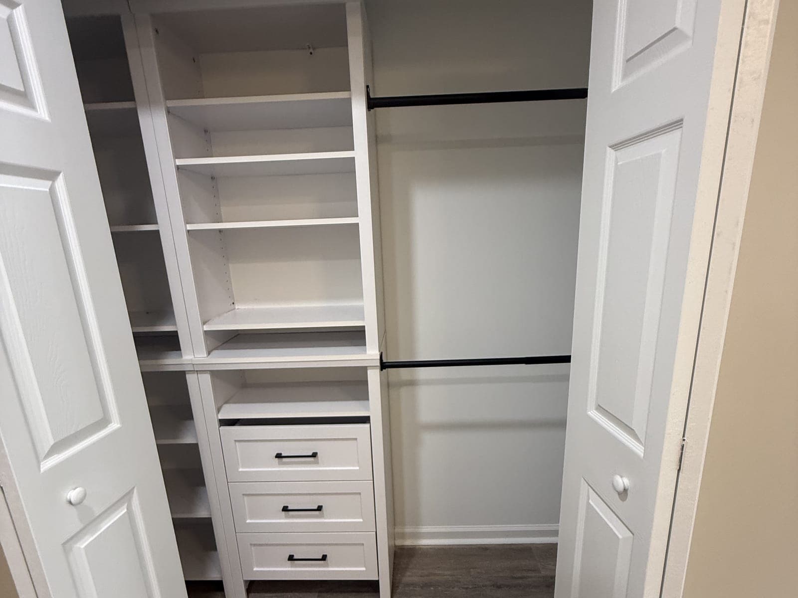 A closet built-in seen through open bifold doors: a white shelving tower with three drawers and black pulls, and two black hanging rods.