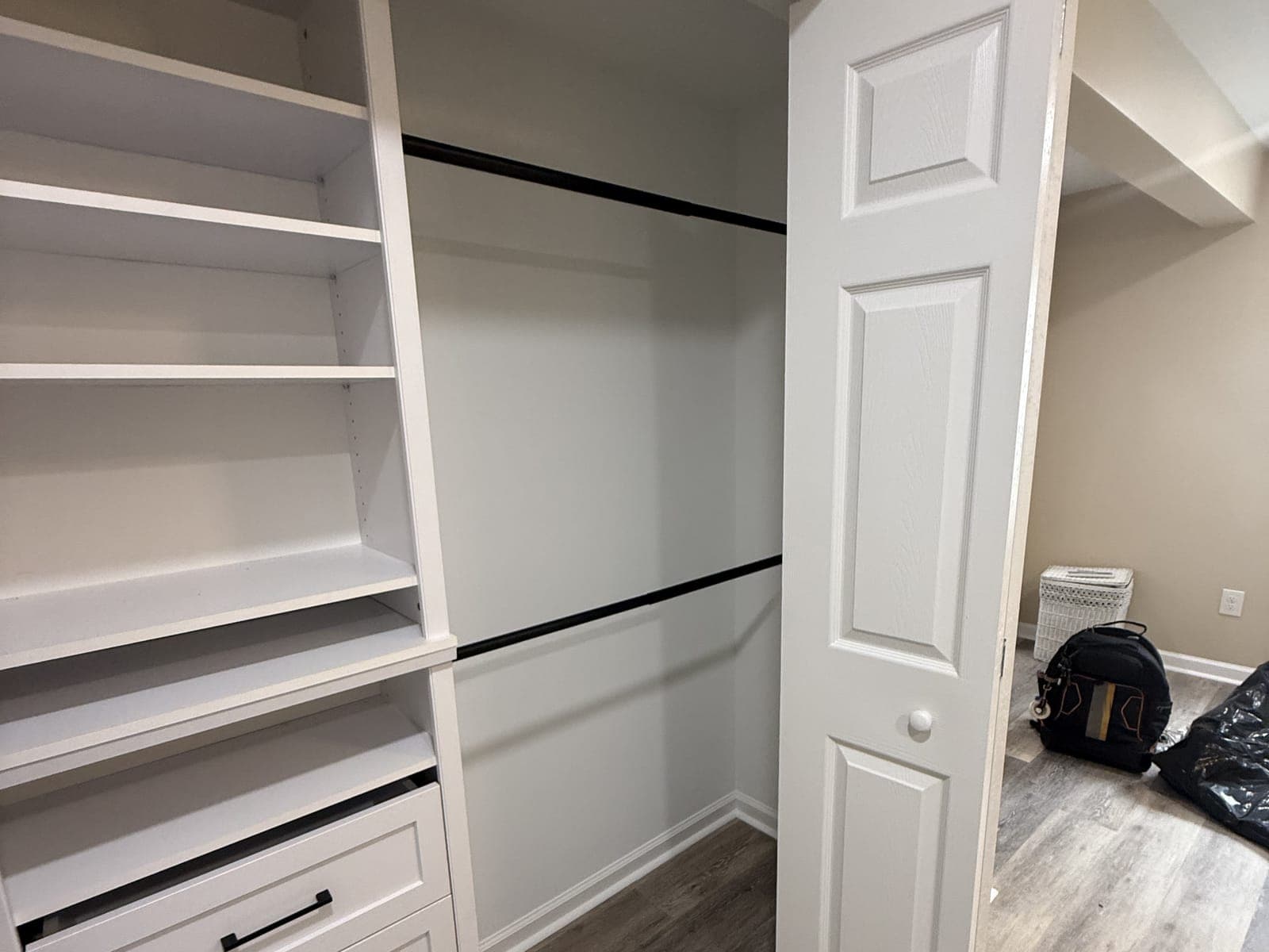 The closet built-in from the side: white shelves and drawers, two black hanging rods, an open bifold door and a plank floor beyond it.