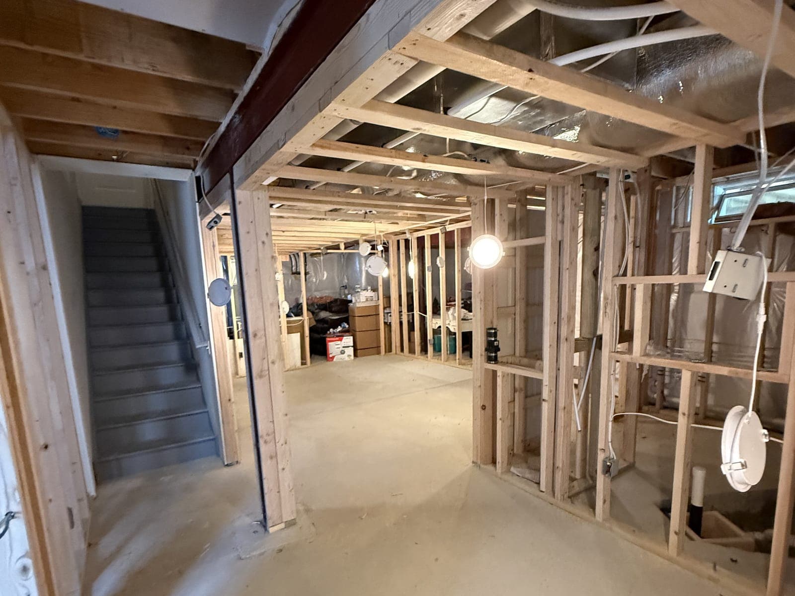 Basement framing from the foot of the stairs: stud walls under the joists, rough wiring, work lights hung from the framing and a long open slab beyond.