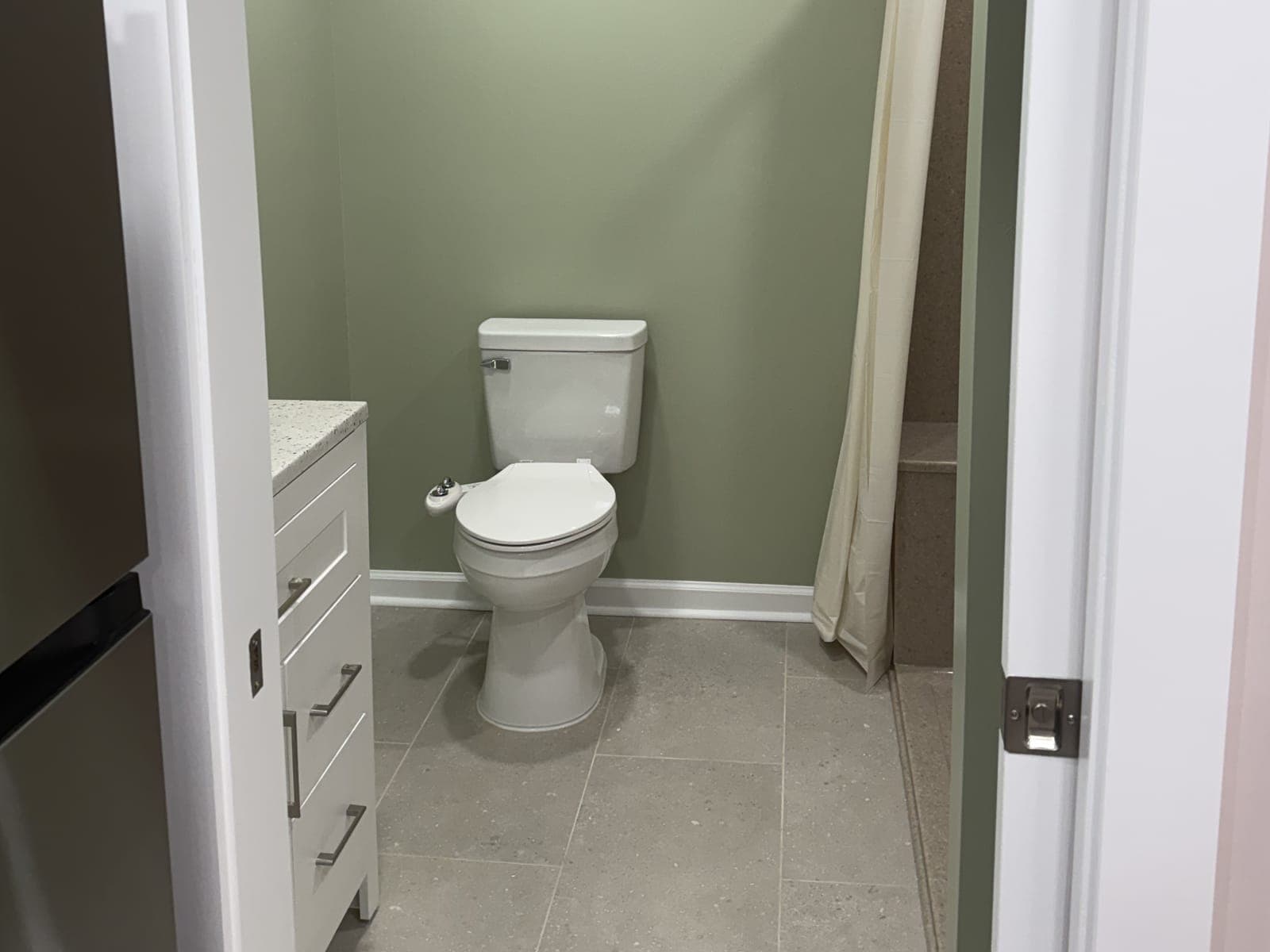 A basement bathroom seen from the doorway: sage walls, a white vanity, a toilet, a gray tile floor and a shower curtain at the right.
