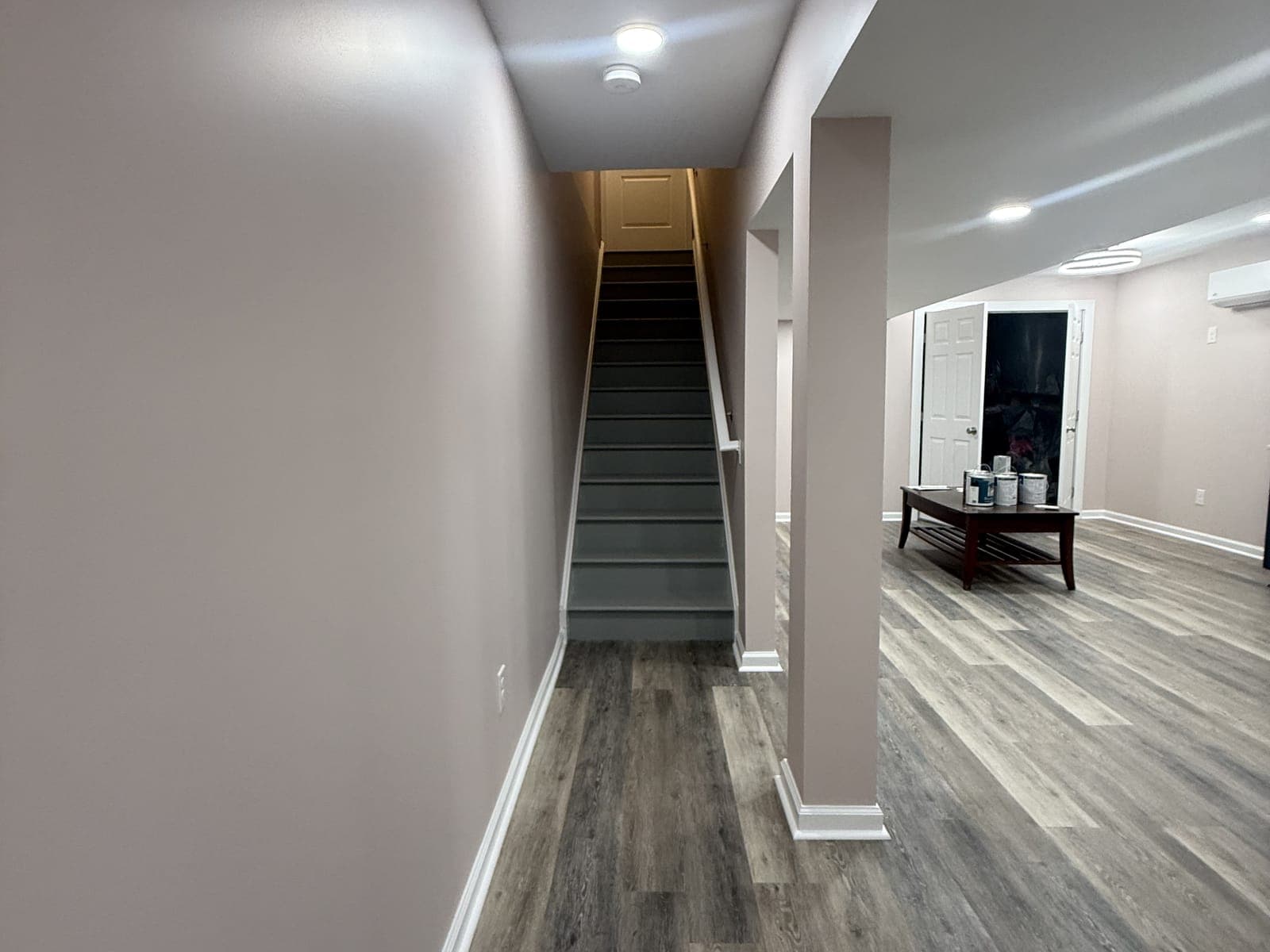 A finished basement hall: painted walls, a plank floor and recessed lights, gray-painted stairs going up at the end, and a wrapped column opening to a room with a coffee table.
