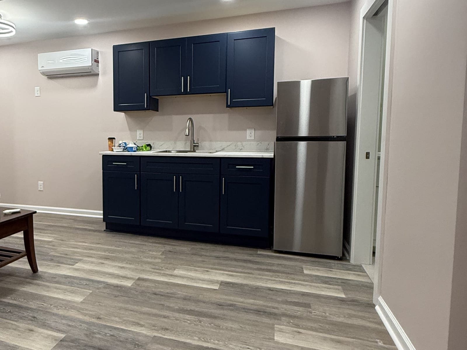 A finished basement kitchenette: navy shaker wall and base cabinets with brushed nickel pulls, a marble-look counter, a stainless sink and a stainless fridge, a mini-split on the wall and a plank floor.