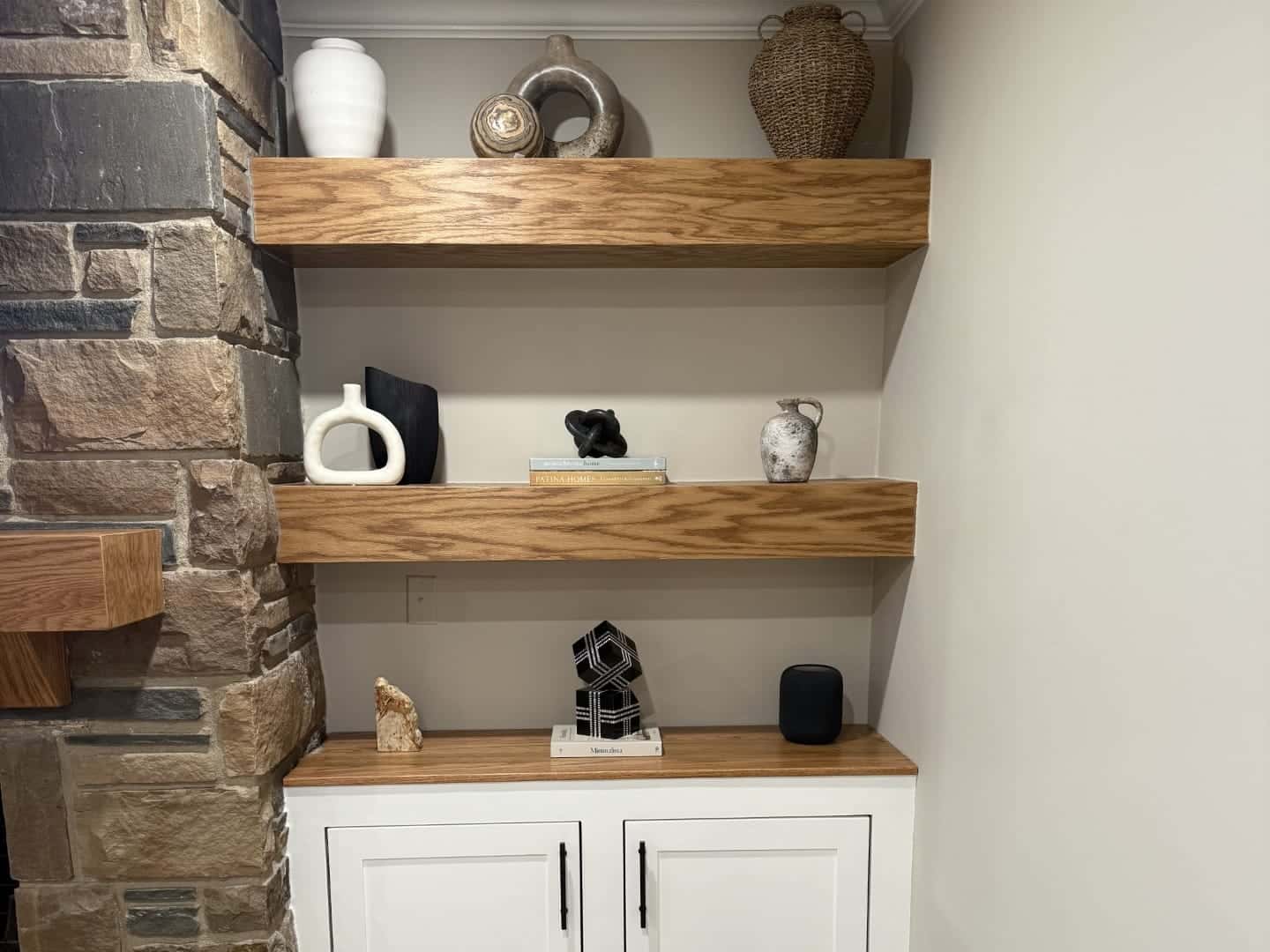 Built-in beside a stone fireplace: three floating oak shelves over a white base cabinet with a wood top and matte black pulls.