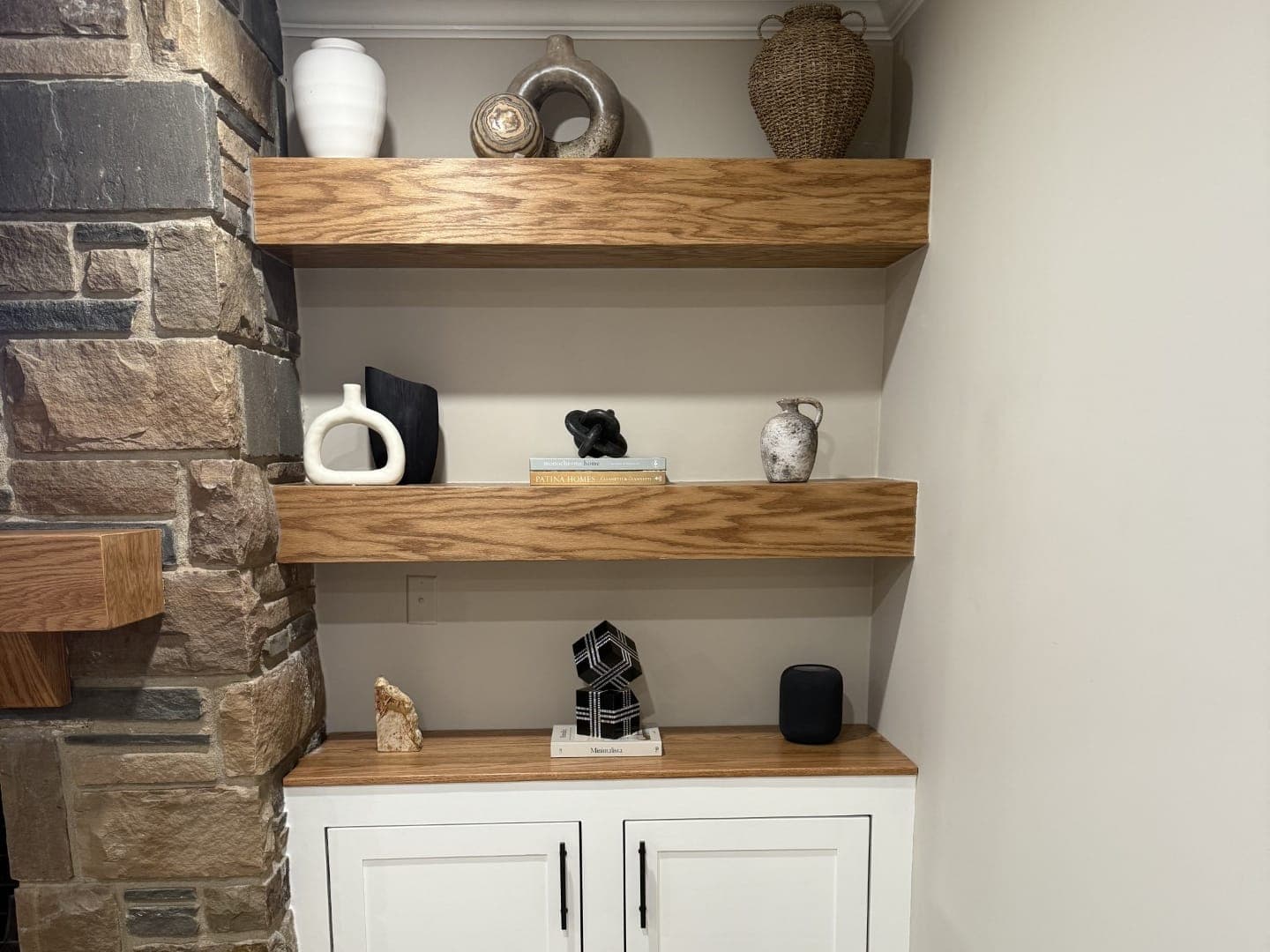 Built-in beside a stone fireplace: three floating oak shelves over a white base cabinet with a wood top and matte black pulls.