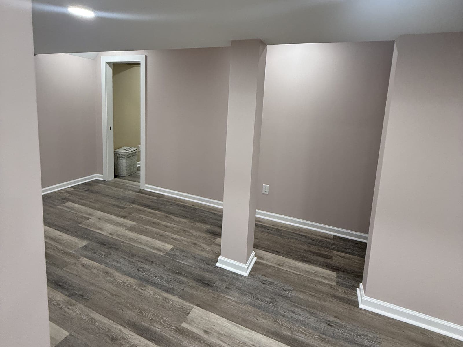 An open finished basement room with a wrapped column, white baseboard, a plank floor and a doorway to a small room with a hamper in it.