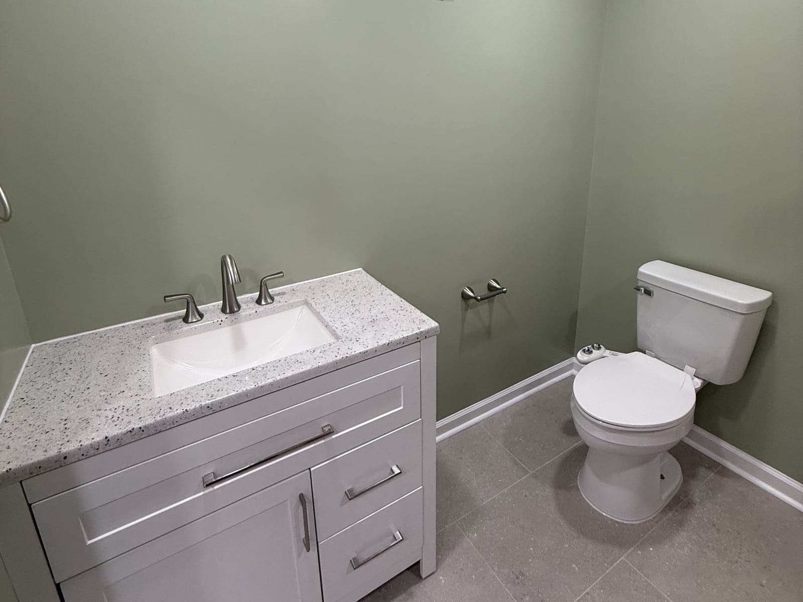 A powder room with sage walls: a white shaker vanity with a speckled top and brushed nickel faucet, a toilet, a paper holder and a gray tile floor.