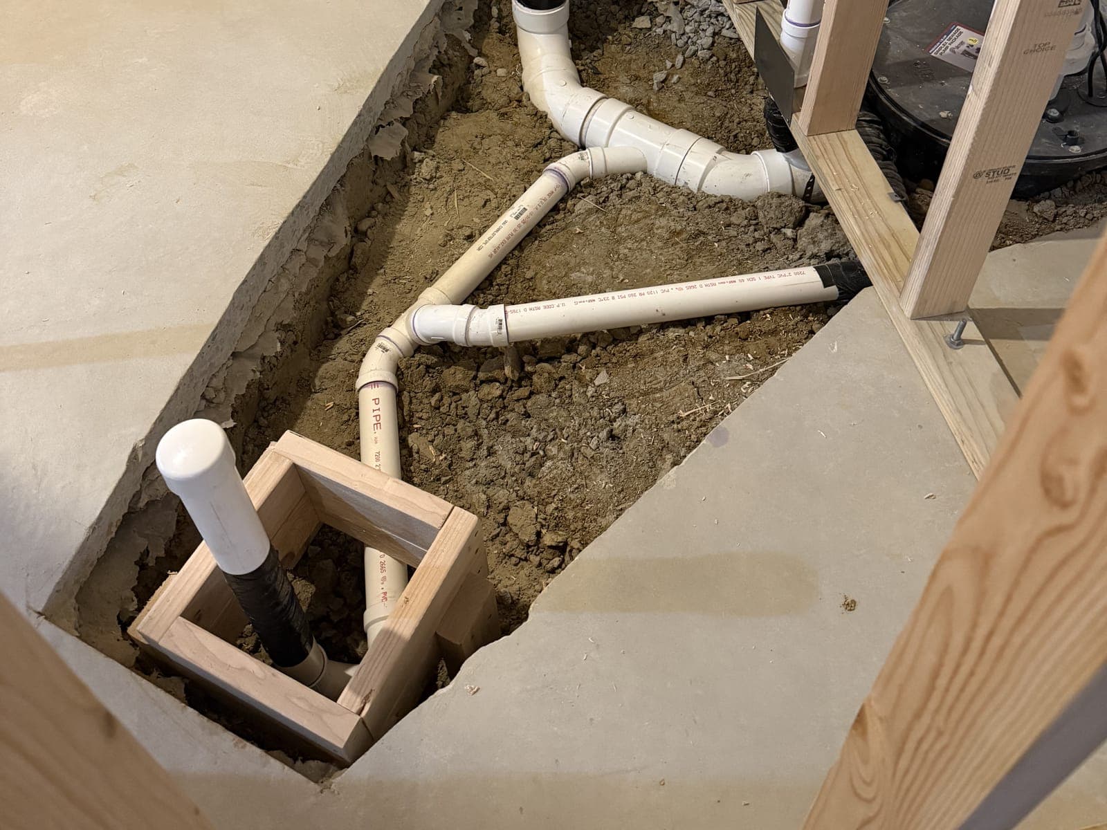 Rough plumbing in a trench cut through a basement slab: white drain pipe and fittings in the dirt, a capped cleanout and a pipe boxed in a wood frame, framed walls behind.
