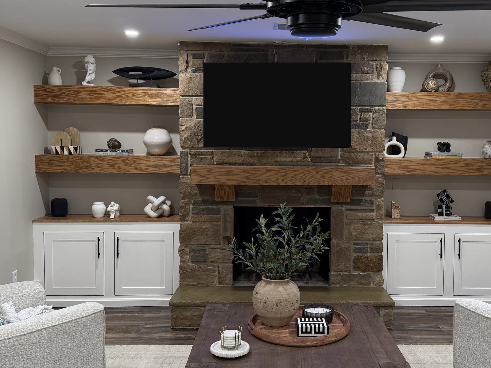 Finished lower-level family room with a stone fireplace, wood mantel, floating oak shelves and white base cabinets.