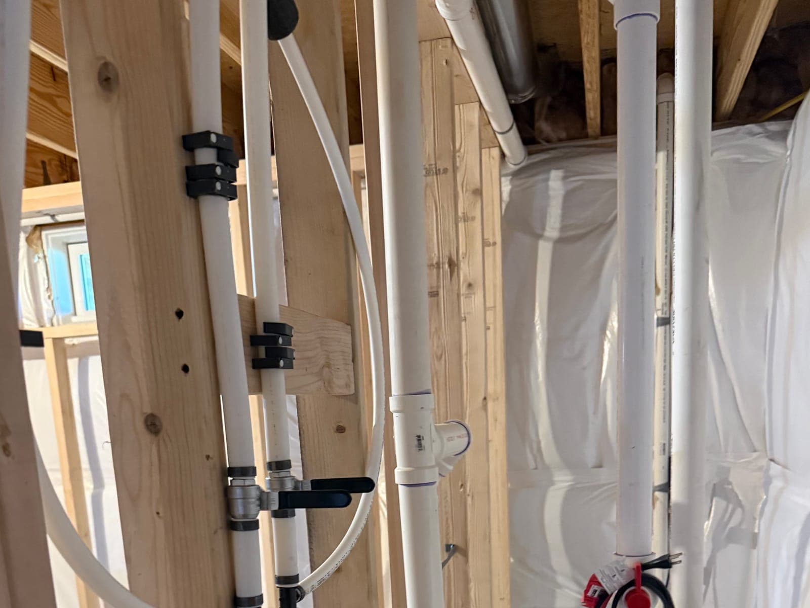 Basement rough plumbing: white supply lines clipped to the studs with two black-handled shut-off valves, a white drain stack and insulated walls behind.