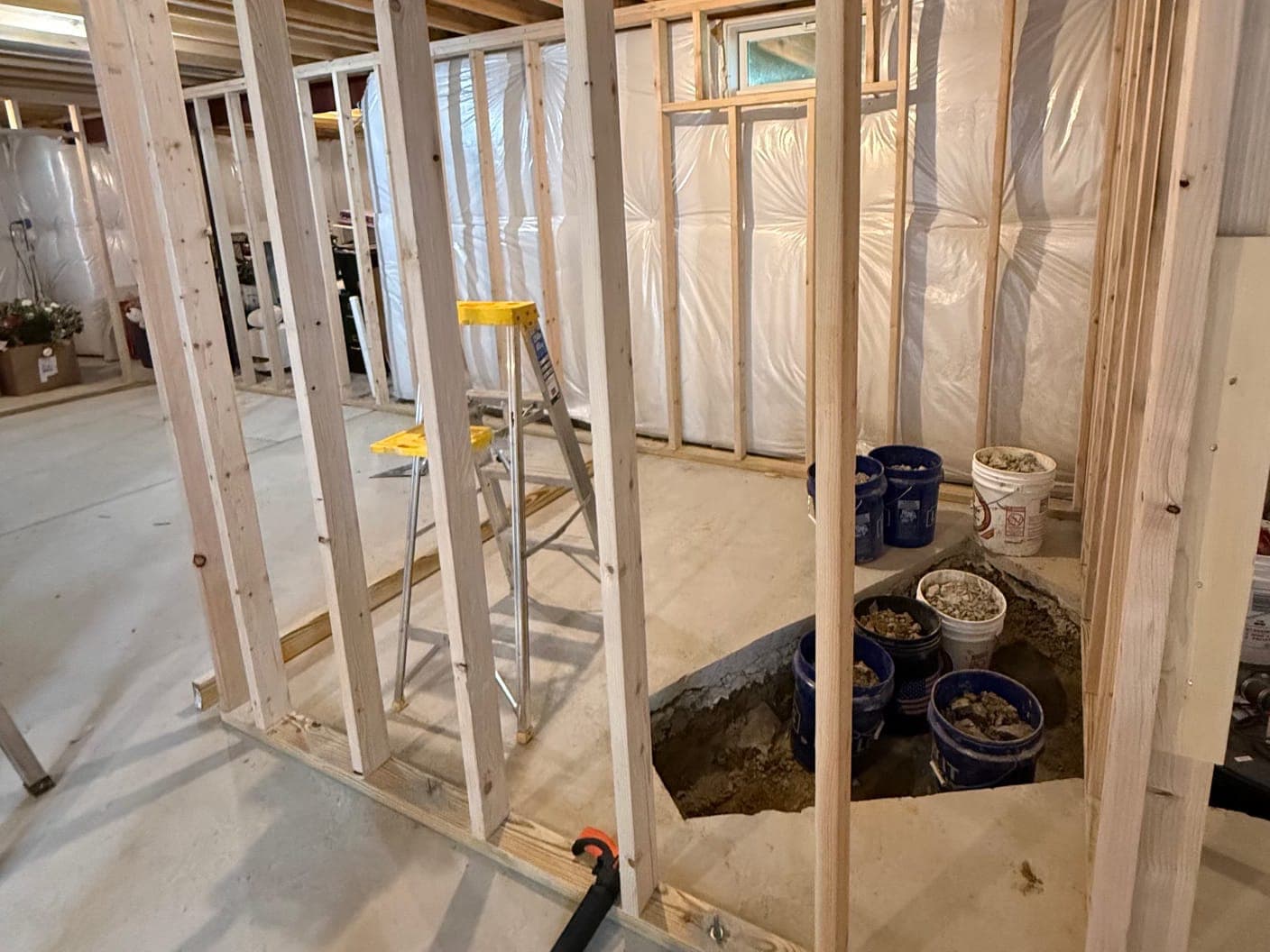 A framed stall in a basement over a trench dug through the slab, buckets of broken concrete in and beside it, a stepladder inside and insulated walls behind.