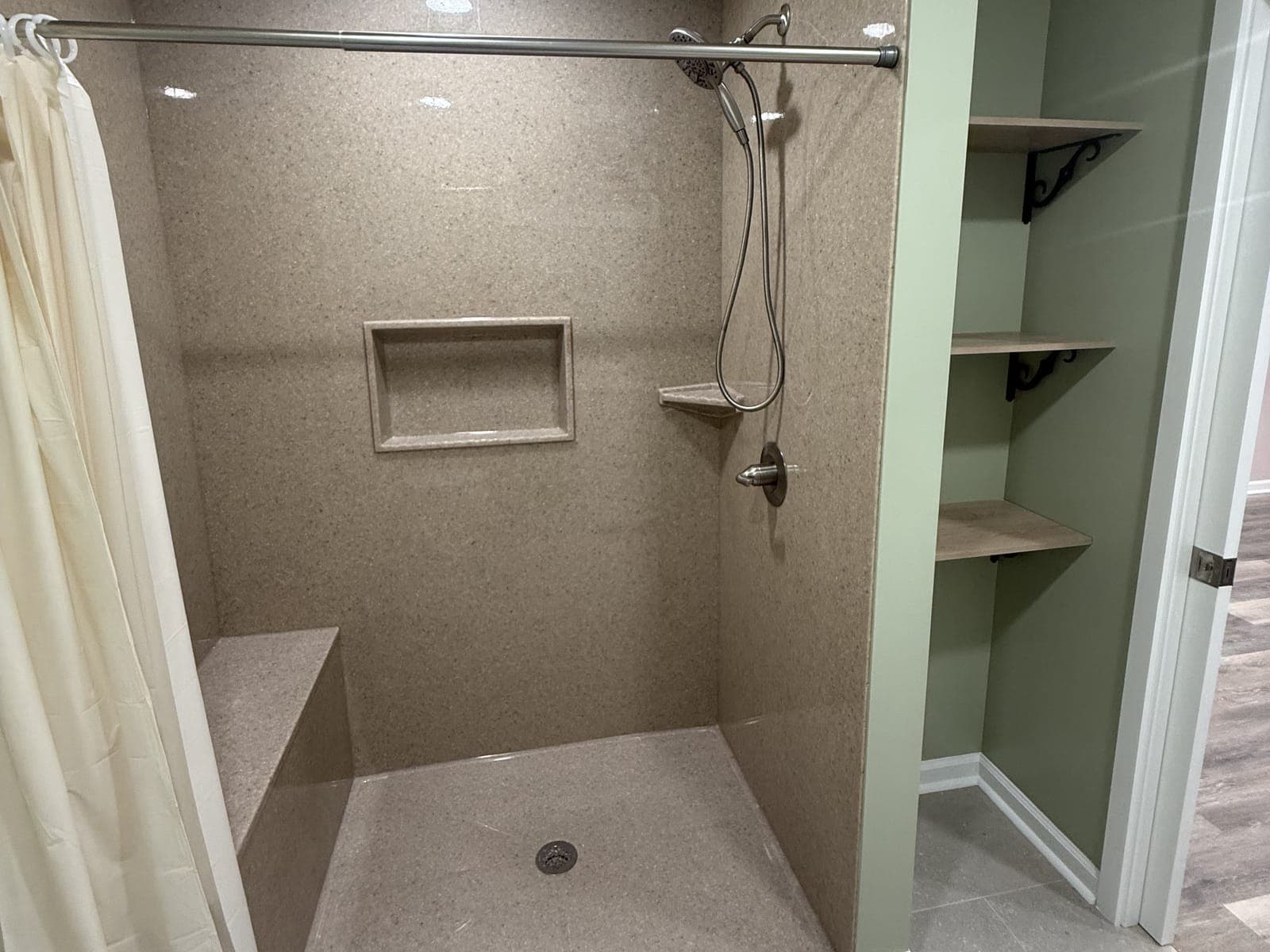 A walk-in shower with solid-surface walls, a molded bench, a recessed niche, a hand shower on a slide bar and a curtain rod, with open shelves on brackets beside it.