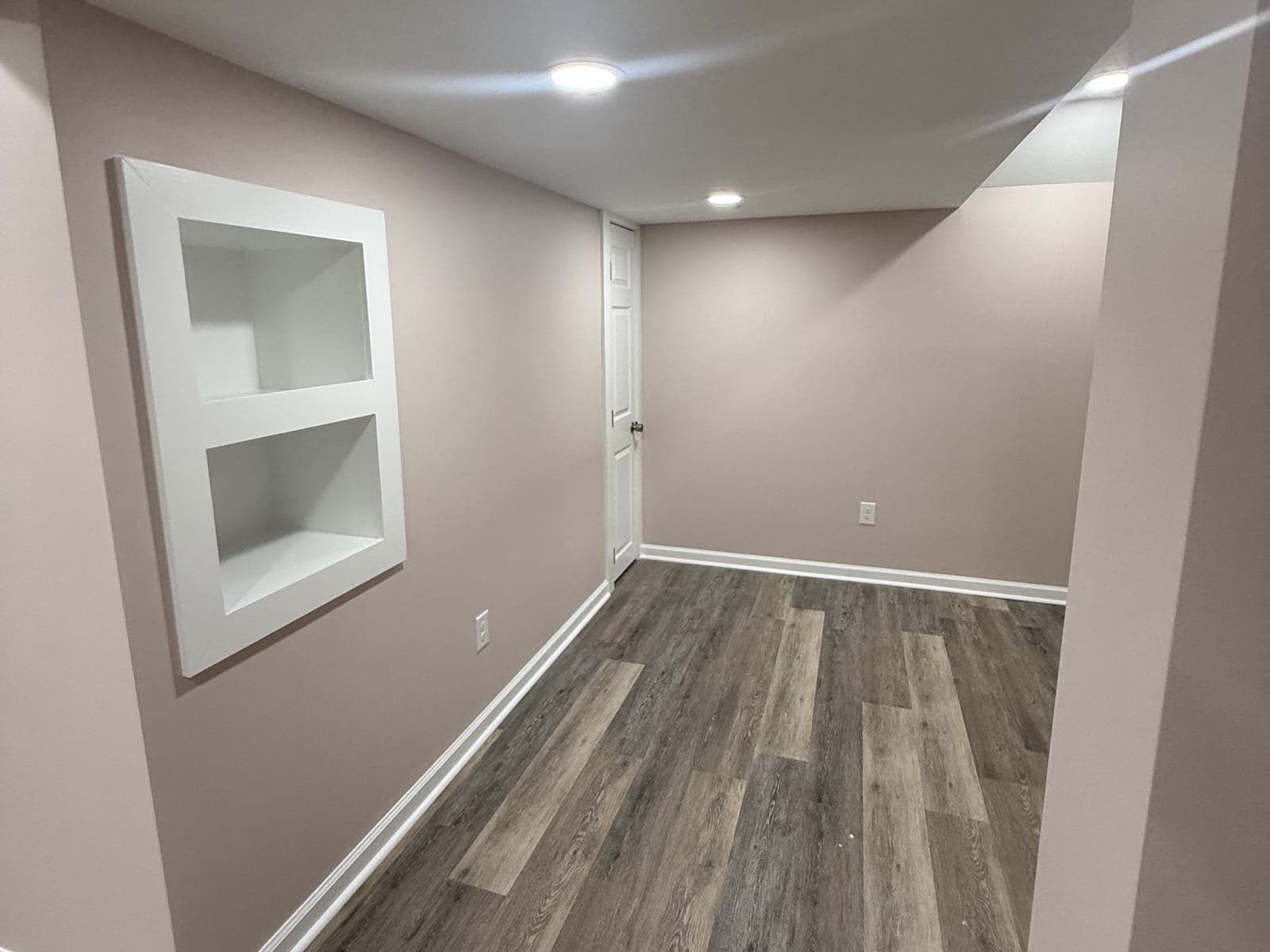 A finished basement room with a recessed two-shelf wall niche in white trim, painted walls, recessed lights, a plank floor and a door at the far end.