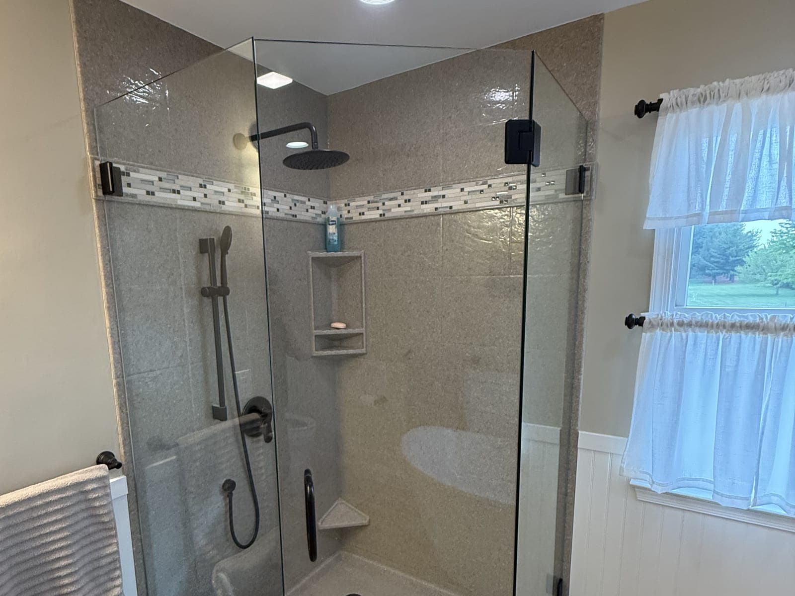 Neo-angle corner shower with solid-surface walls, a glass mosaic band, matte black fixtures and a frameless glass enclosure.