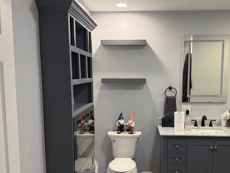 Bathroom with a tall charcoal built-in cabinet, gray floating shelves, a gray vanity with a marble-look top and black faucet, and a lit mirror.