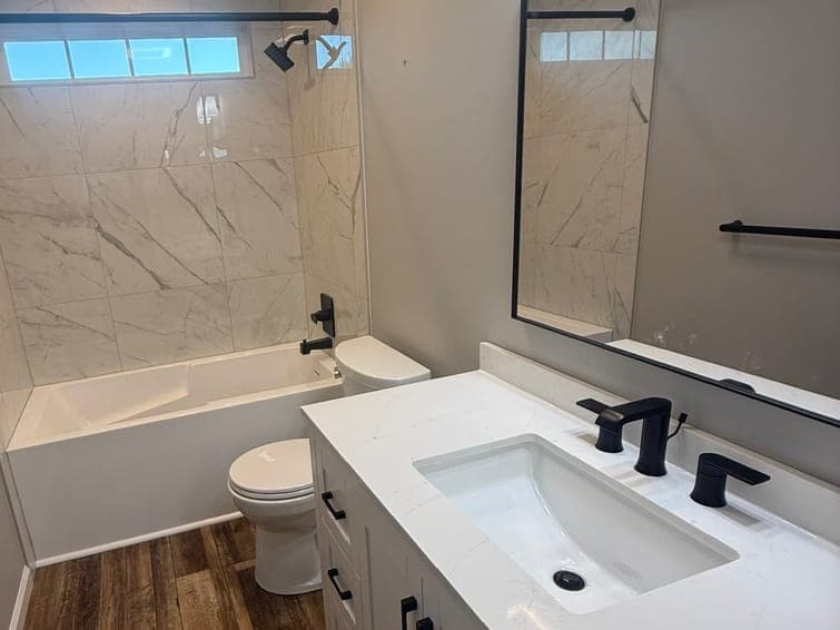 Bathroom with a marble-look tile tub surround and a glass block window, a white shaker vanity with a white top, a matte black faucet and towel bar, and a wood-look plank floor.