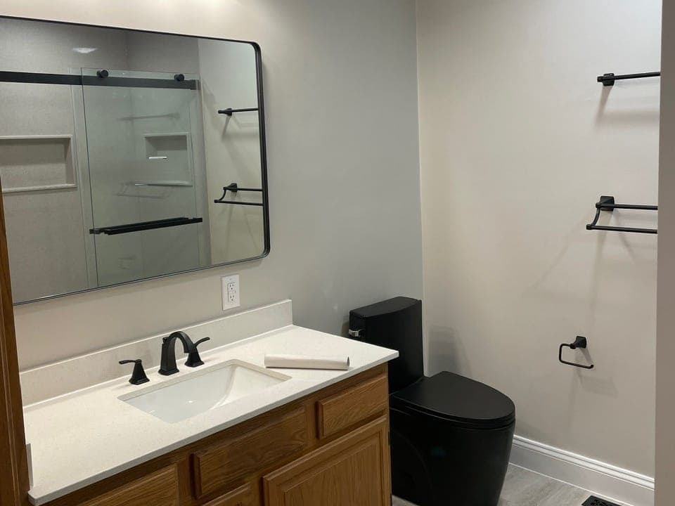 Bathroom with an oak vanity and white top, a matte black faucet, a black-framed mirror that shows the shower's sliding glass door, a black one-piece toilet and black towel bars.
