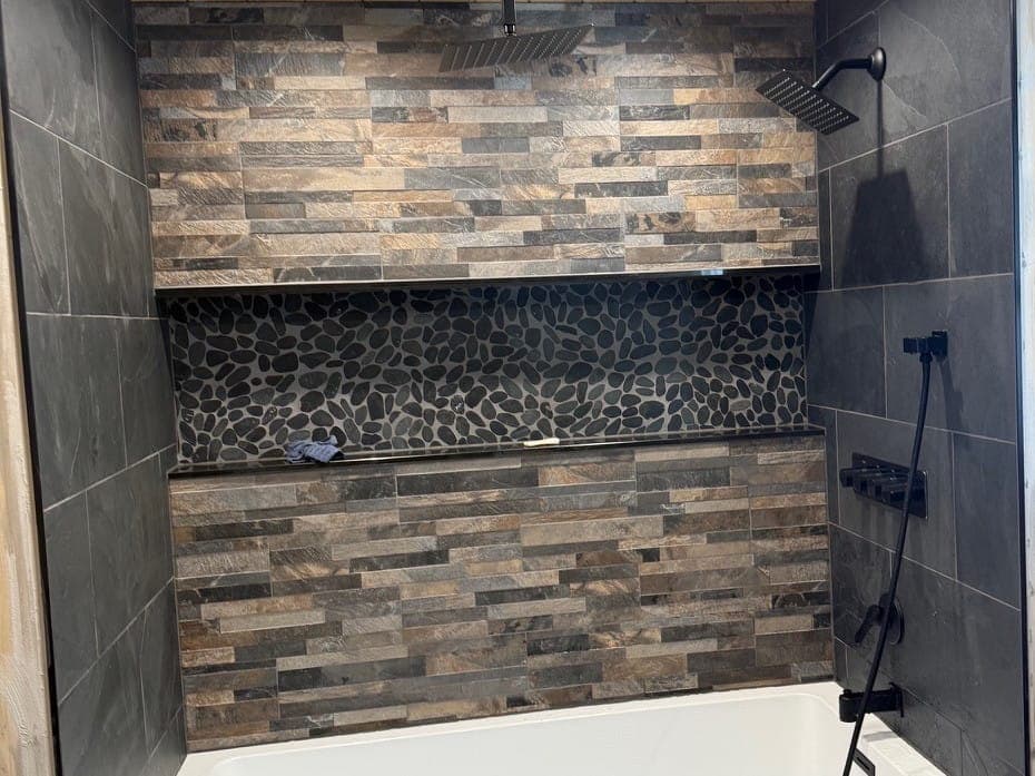 Tub surround mid-build: a knotty pine ceiling with recessed lights, a rain shower head, stone-look ledger tile with a pebble niche, dark slate-look tile and matte black fixtures over a white tub.