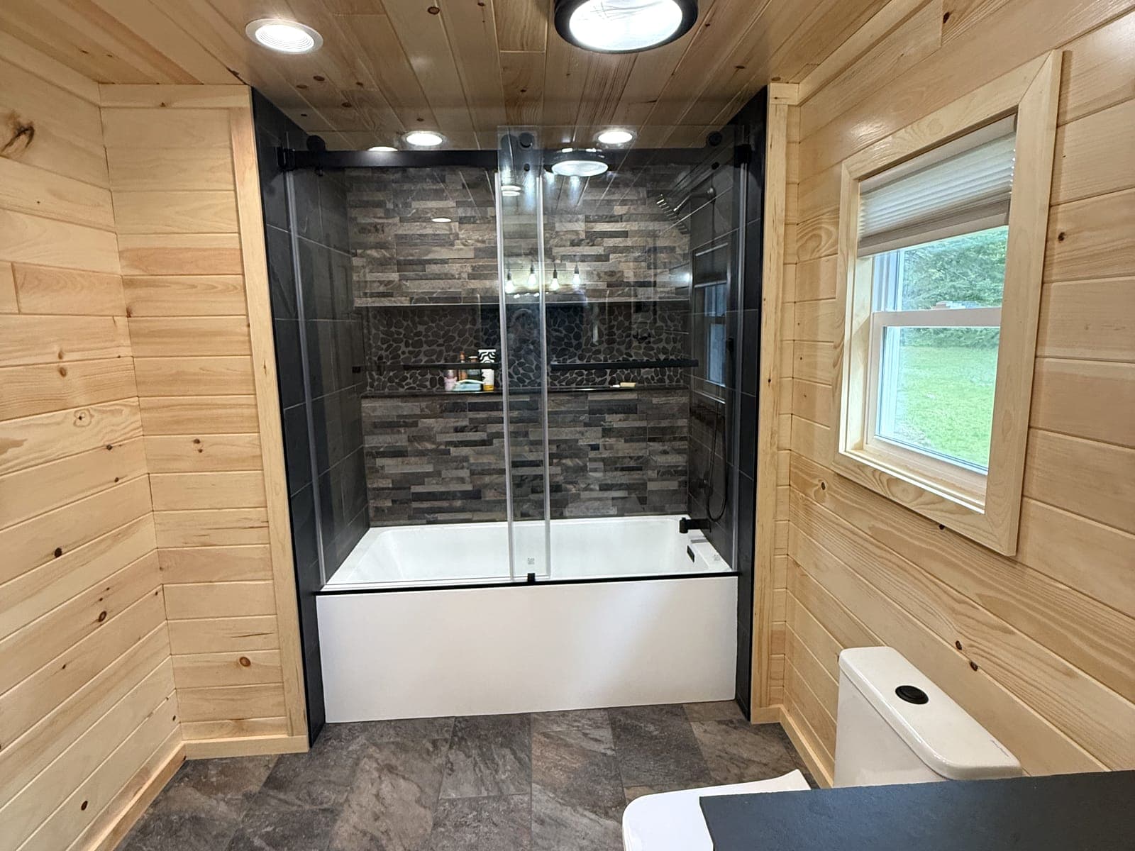 Bathroom with knotty pine walls and ceiling, a tub and shower in dark slate-look tile with a pebble niche, and a black-framed sliding glass door.