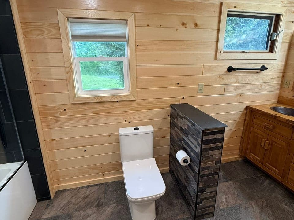 Knotty pine bathroom wall with two small windows in pine trim, a white toilet, a tiled half-wall beside it and a log vanity at the edge.