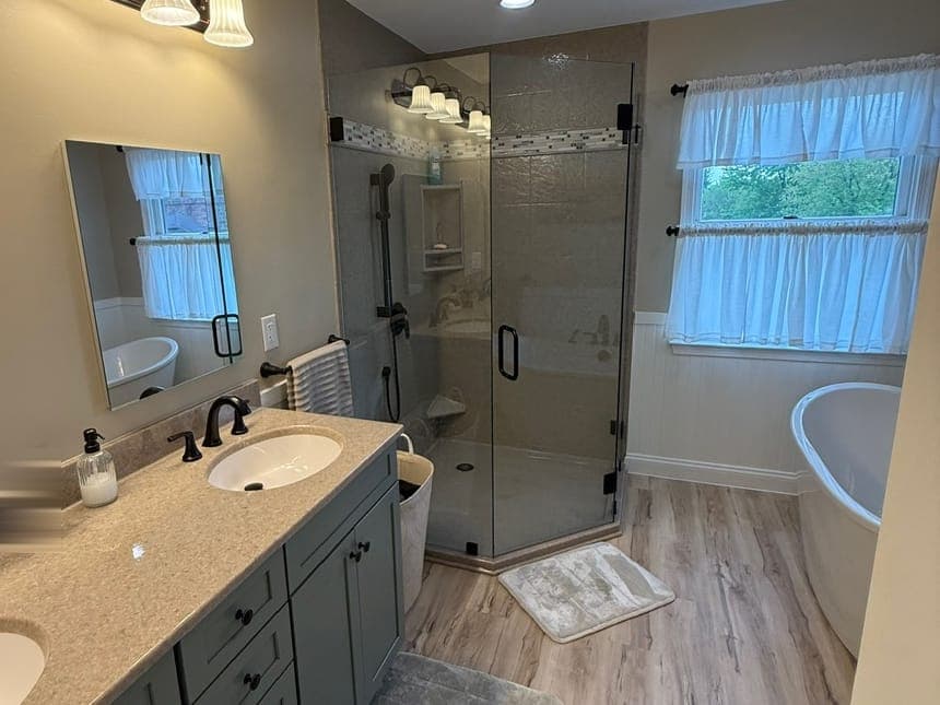 Bathroom with a sage double vanity, a neo-angle glass corner shower, a freestanding tub under a curtained window and a wood-look plank floor.