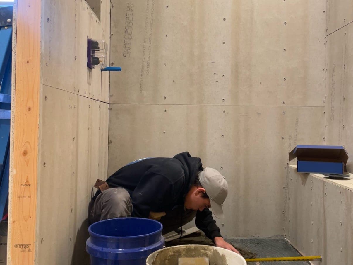 A shower mid-build: cement board walls with the valve and niche framed in, and Dominic Dawson in a cap and hoodie packing the mortar bed over metal lath, a tape measure across the floor and buckets in front.