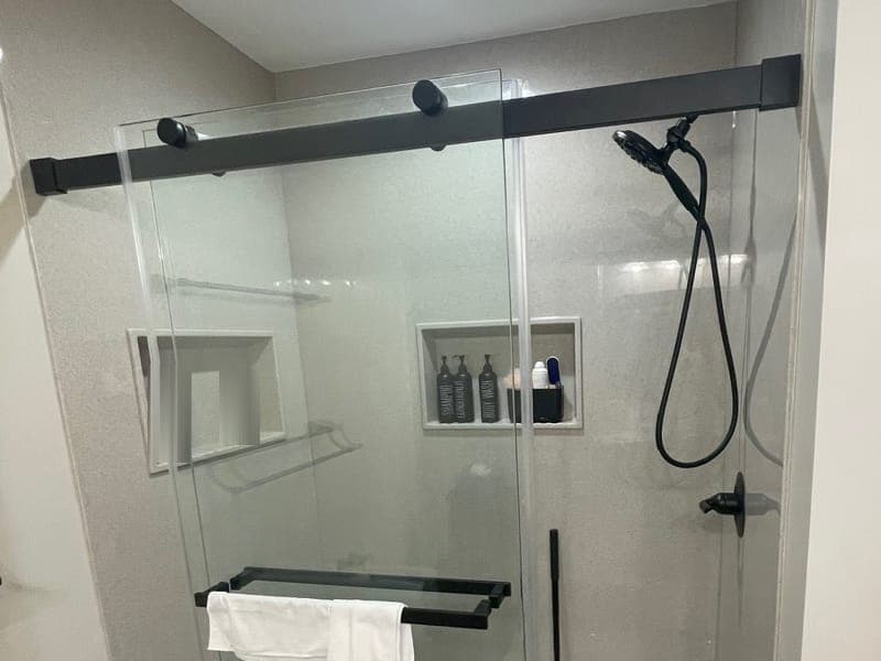 Shower with solid-surface walls, two niches, a matte black hand shower and a black-framed sliding glass door with a towel over its bar.