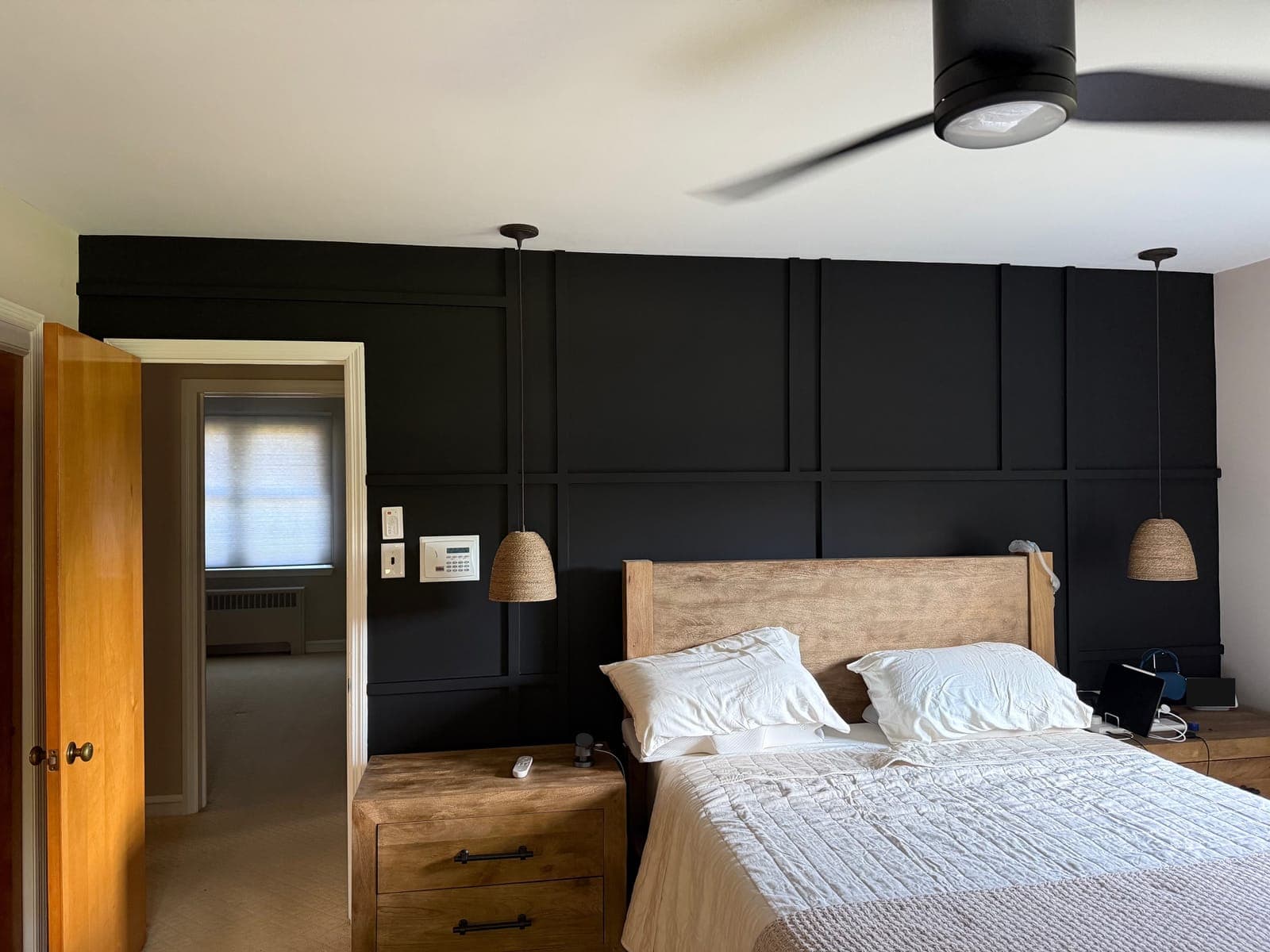 A bedroom accent wall with a grid of battens, all painted black, two woven pendants hung on either side of a wood bed, and a ceiling fan above.