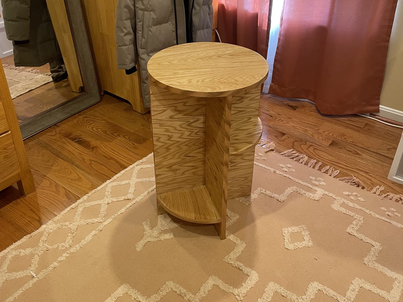 A round oak side table with a half-round lower shelf on a patterned rug, a wardrobe and a floor mirror behind it.