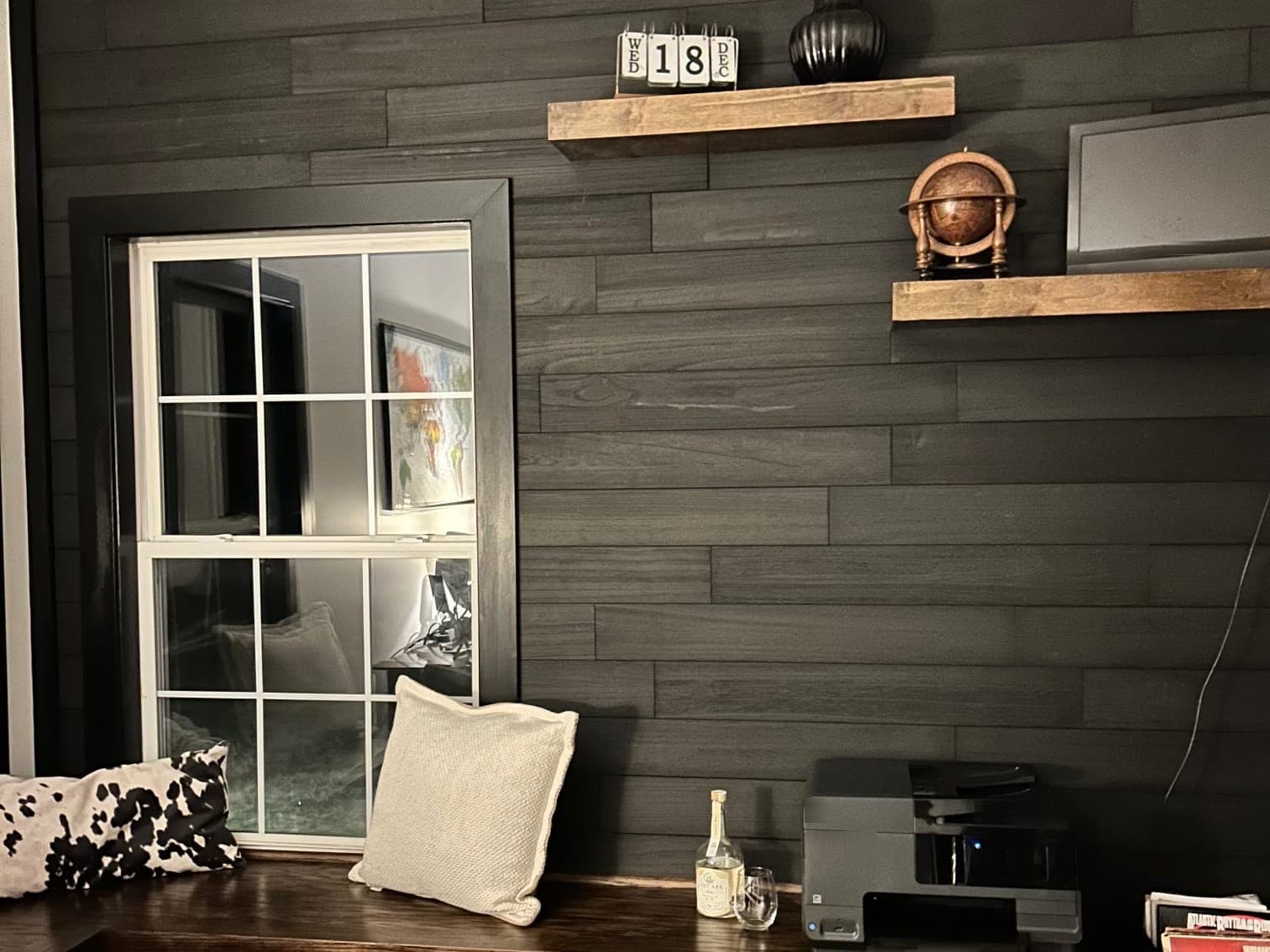 A dark stained plank wall with two stained floating shelves, a wide black casing around a window, and a stained bench built along the wall with pillows, a printer and a record crate on it.