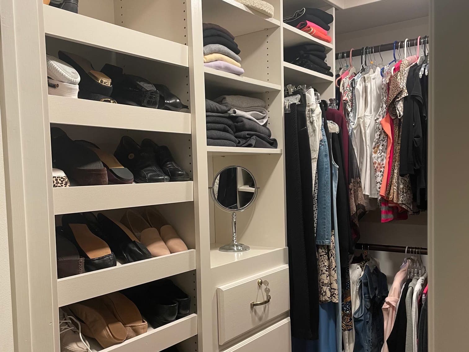 A white closet built-in in use: shoes on sloped shelves, folded sweaters in cubbies, a vanity mirror over a drawer bank, and clothes on two rods.