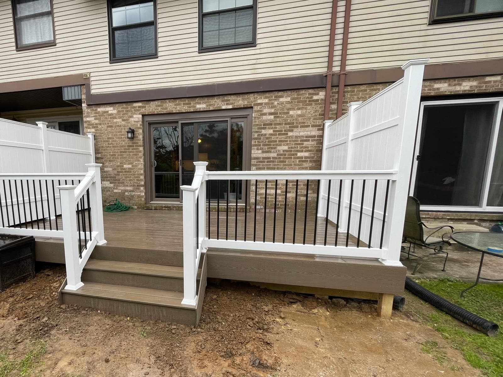 A finished composite deck seen from the yard: steps, white rails with black balusters and white privacy fence panels against a brick and siding house.