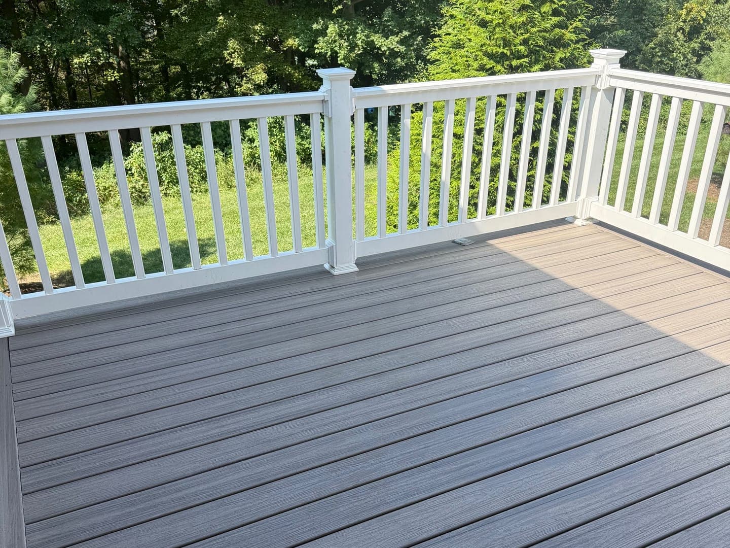 A finished deck with gray composite boards, a border board along the edge and a white rail with white balusters, trees and a lawn beyond.