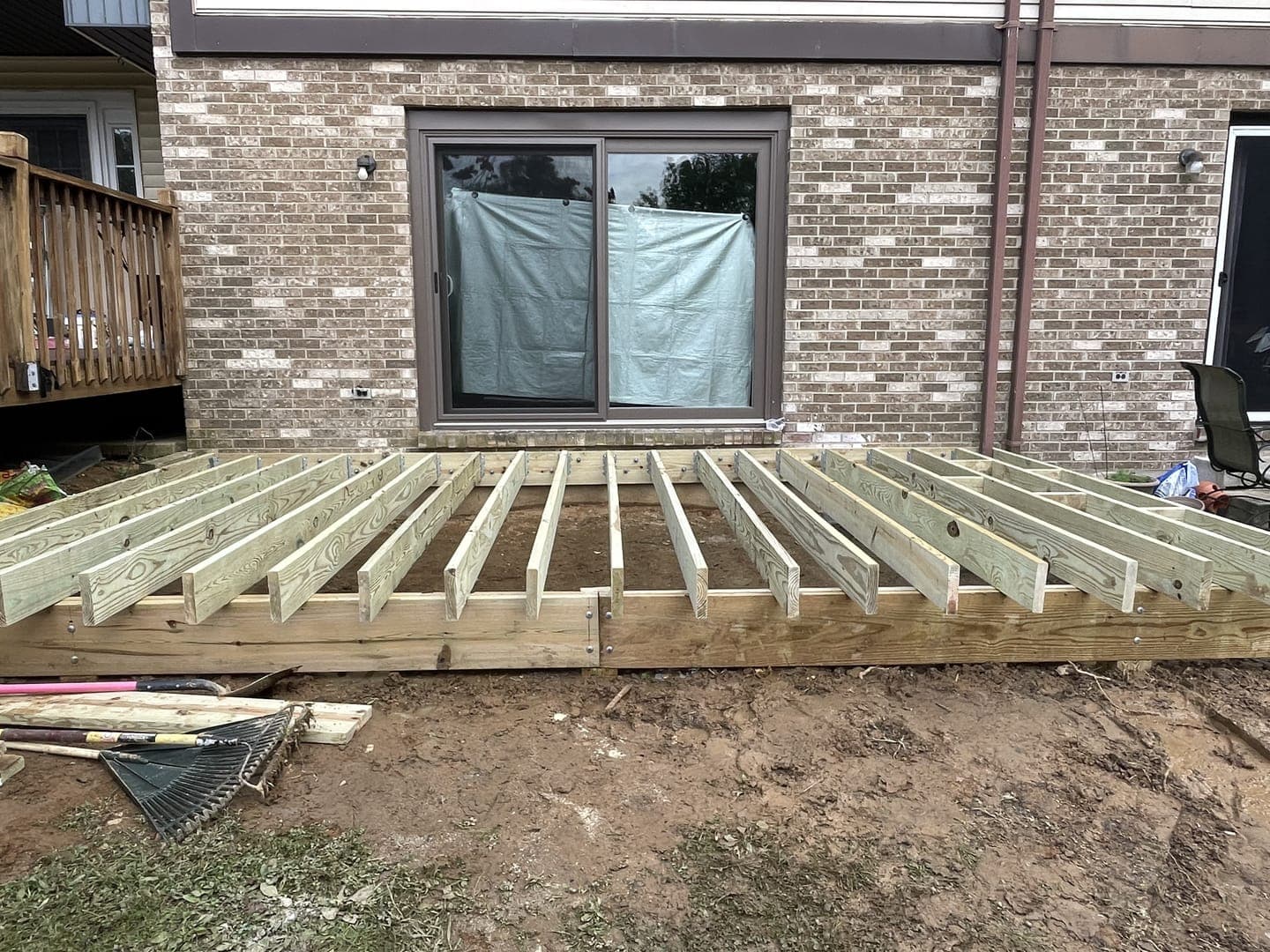 Deck framing against a brick house: pressure-treated joists on a rim board, a covered sliding door behind, and bare dirt in front.