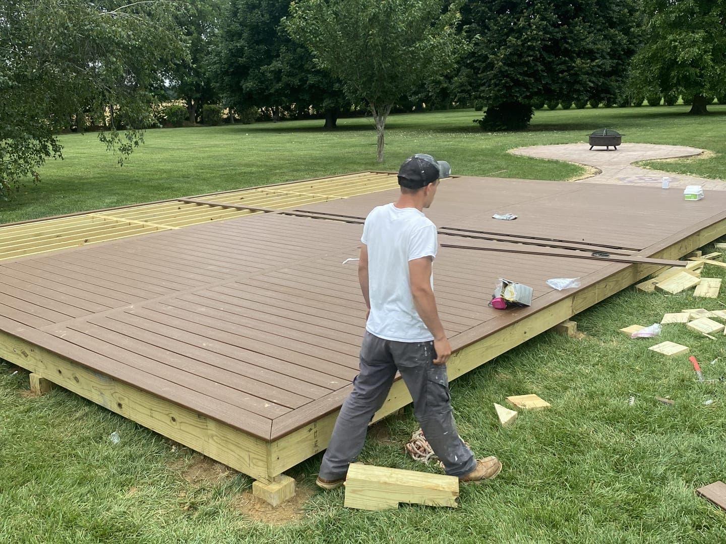 A freestanding deck mid-build in a lawn: brown composite boards with a border, open joists at the far end, and a person walking beside it.