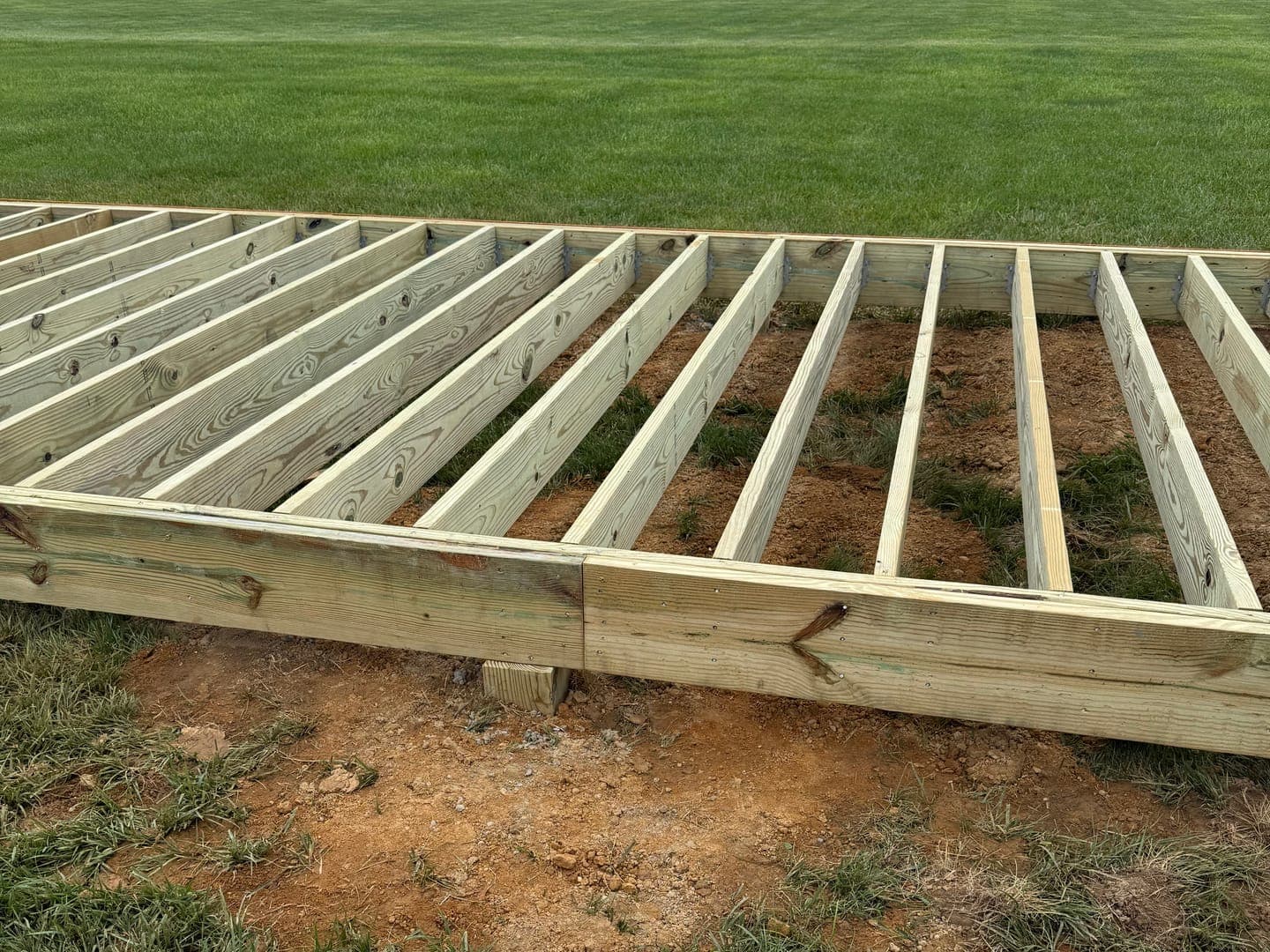 Ground-level deck framing in a lawn: pressure-treated joists in hangers inside a pressure-treated rim, on a block over bare dirt.