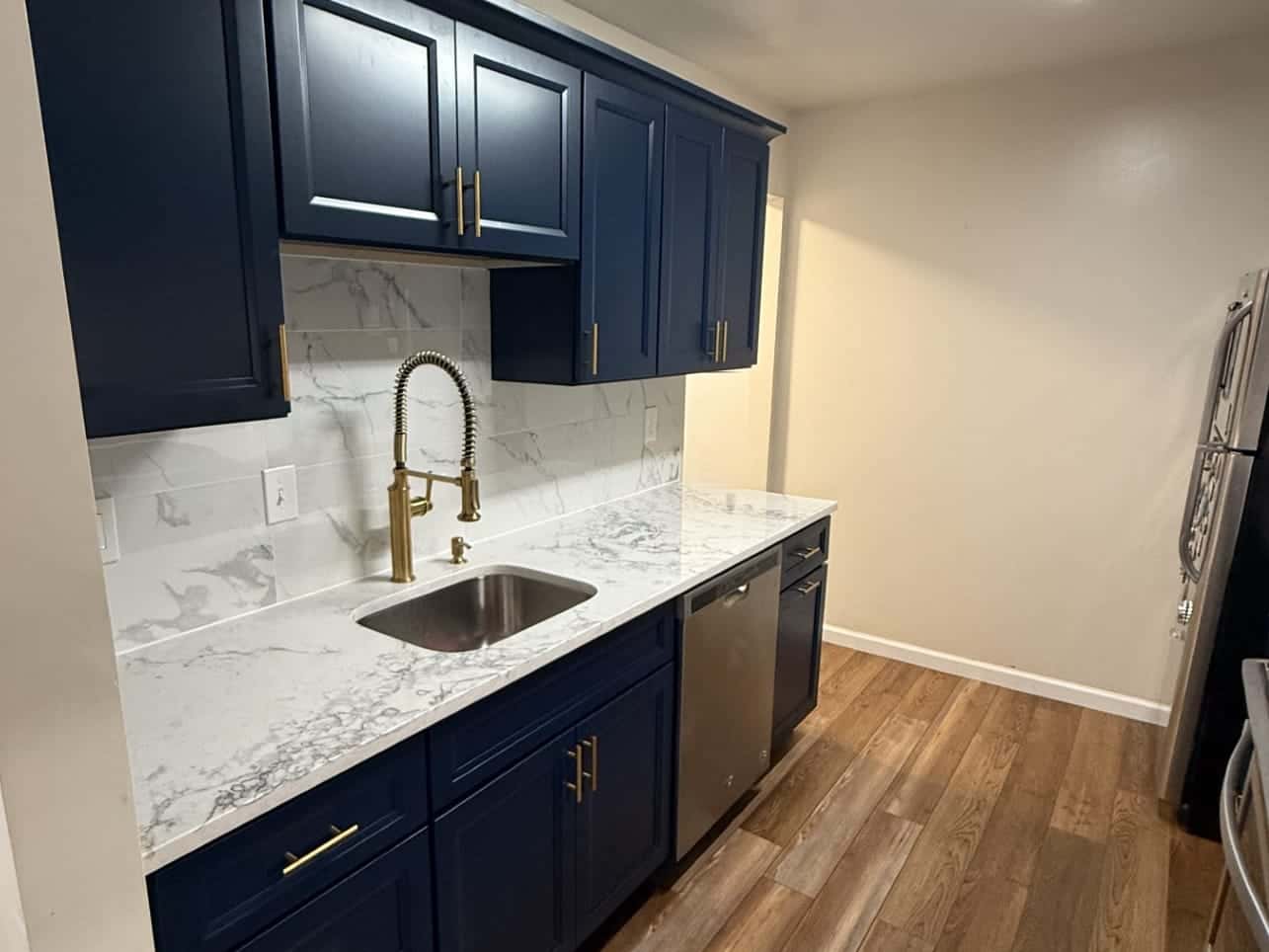 Galley kitchen sink wall with navy cabinets, brushed gold pulls, a gold spring-neck faucet, an undermount stainless sink, a marble-look counter and a marble-look tile backsplash.