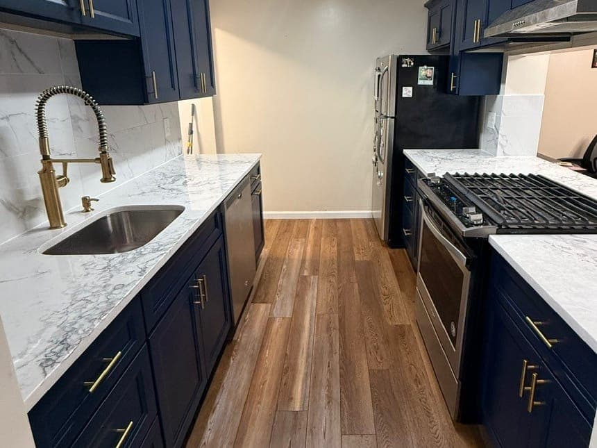 Galley kitchen straight down the middle: navy cabinets with gold pulls on both sides, a marble-look counter, a stainless gas range and a wood-look plank floor.