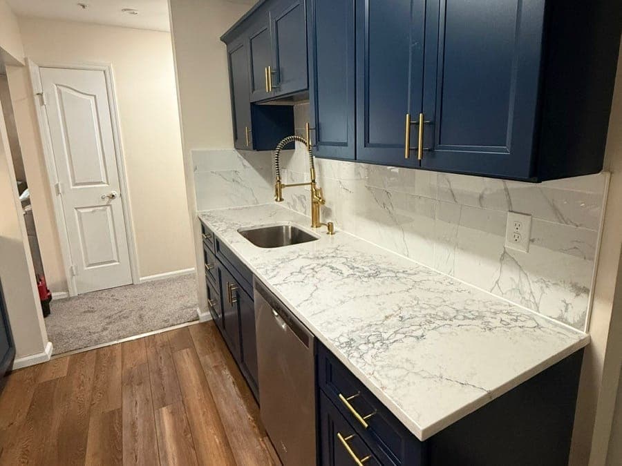 Galley kitchen sink wall seen from the hall: navy cabinets with gold pulls, a gold spring-neck faucet, a marble-look counter and backsplash, and a wood-look plank floor.