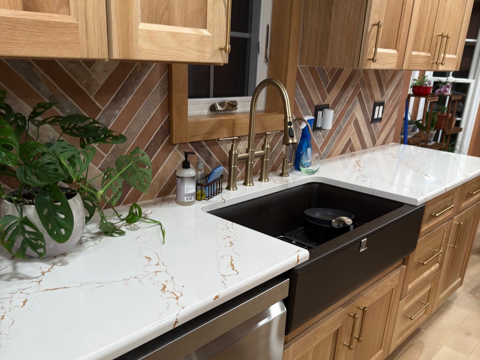 Kitchen sink run with oak cabinets and gold bar pulls, a black farmhouse sink, a brushed gold bridge faucet, a white counter with gold veining and a herringbone tile backsplash around a window.