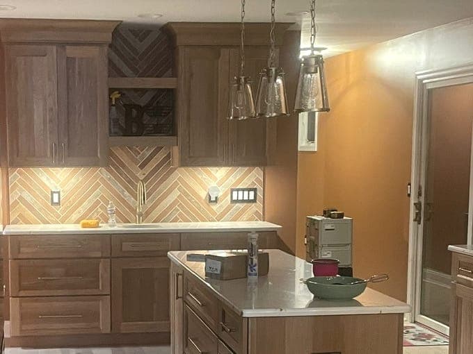 Kitchen with oak cabinets, a herringbone tile backsplash, an open shelf over the range wall, three glass pendants over an island with a white top, and a plank floor.
