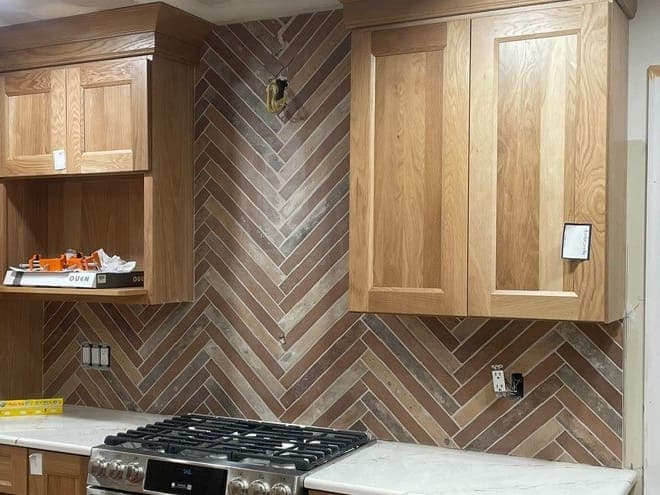 Kitchen range wall with oak cabinets and crown molding, a stainless gas range and a herringbone tile backsplash in brown and gray.